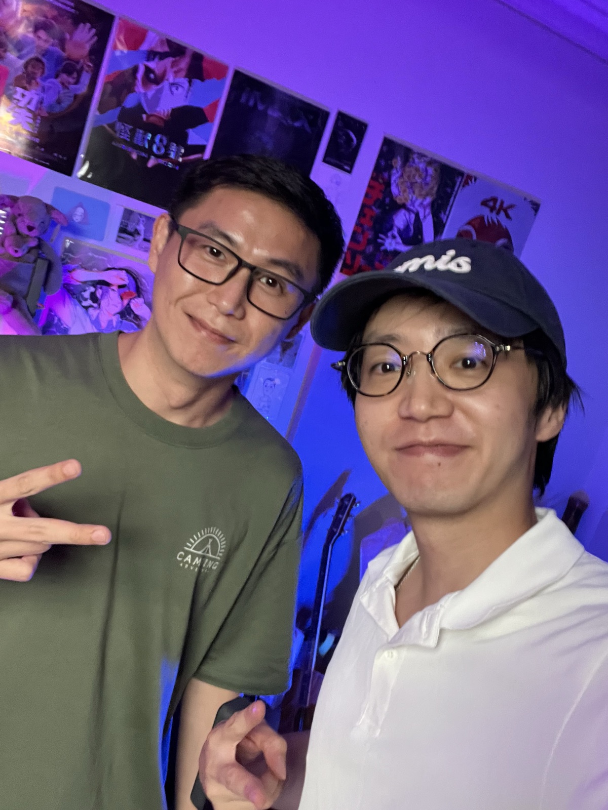 藍紫燈光的工作室裡兩個人並肩合照，其中一人比出手勢，背景是牆上的海報。