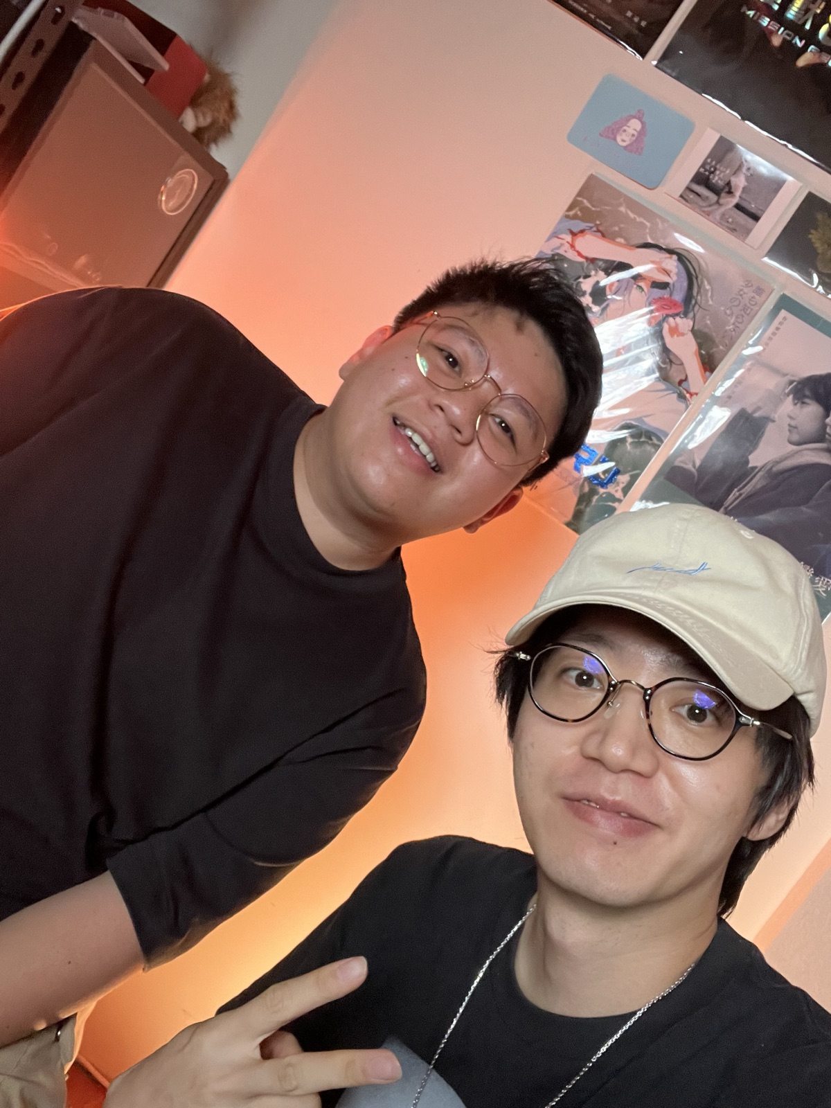 暖橘燈光的工作室裡兩個人臉靠著臉看向鏡頭合照，牆上貼著海報。